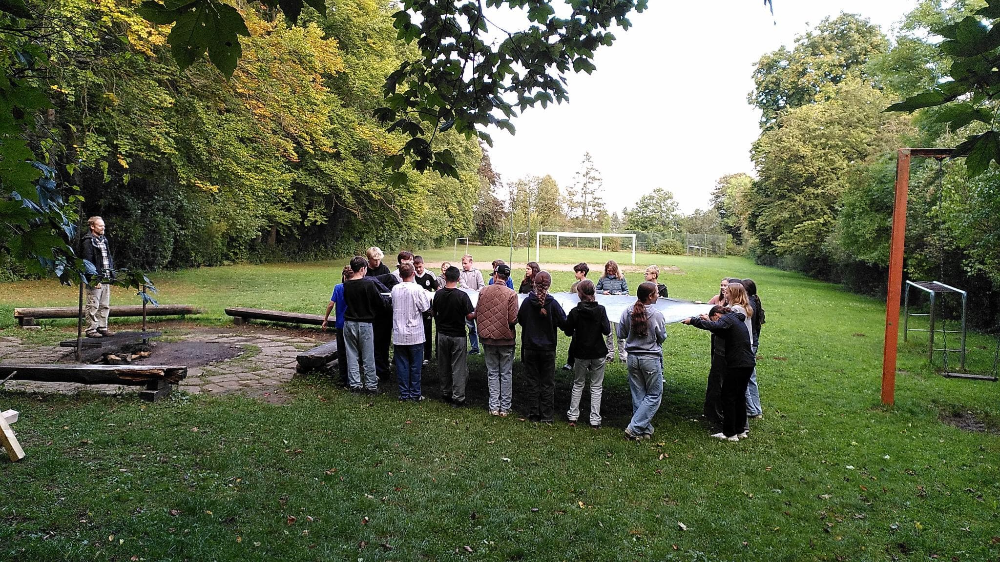 Eine Klasse steht mit ihren Teamer:innen im Kreis auf einer Wiese